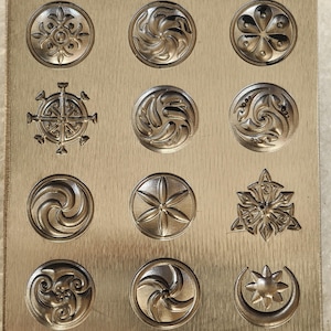 May include: A metal plate featuring twelve circular designs with geometric patterns. The designs include a compass rose, floral patterns, and swirling motifs. The plate has a brushed metal finish, possibly for crafting or decorative use.