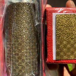 以下が含まれることがあります： エンボス加工された渦巻き模様の装飾的な円筒形のオブジェクトのクローズアップ。プラスチックに包まれています。その隣には、金色のテクスチャ加工された表面を持つ小さな赤い箱があります。アイテムは手で持たれています。