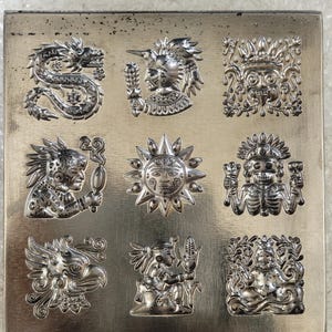 May include: A square silver-toned metal plate featuring a grid of nine embossed designs. The motifs include a coiled dragon, a warrior figure, a decorative mask, a profile of a person, a sunburst, a skeleton figure, an eagle, a person holding corn, and a seated deity.