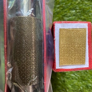 以下が含まれることがあります： 複雑な模様の装飾用金属オブジェクトで、赤い箱に入っています。オブジェクトは銀色のベースと、テクスチャ加工されたブロンズ色のトップを備えています。小さな金色の模様の正方形も表示されています。