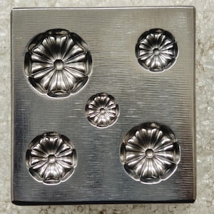 May include: A square metal dapping block with a brushed finish featuring five circular indentations of varying sizes. Each indentation has a radial, flower-like pattern embossed into the surface, designed for shaping metal components.