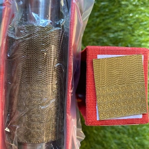 Puede incluir: Un tapón de botella decorativo, metálico, con un intrincado patrón en relieve, presentado en una caja roja. El tapón está envuelto en plástico transparente, y una tarjeta dorada con un diseño a juego es visible.
