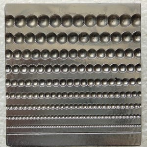 Könnte beinhalten: Ein quadratischer Metall-Dapping-Block mit einer Reihe horizontaler Linien, die halbkugelförmige Vertiefungen unterschiedlicher Größe aufweisen, von großen kreisförmigen Mulden oben bis zu einer Reihe winziger perlenartiger Abdrücke unten.