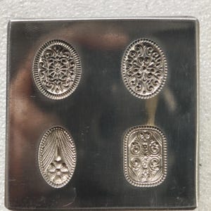 Puede incluir: Una herramienta cuadrada de metal plateado con cuatro diseños decorativos en relieve. Los diseños incluyen dos formas ovaladas y otras dos formas, cada una con intrincados patrones florales y de volutas. La herramienta parece ser utilizada para la fabricación de joyas o el trabajo de metales.