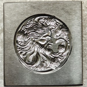 Puede incluir: Una placa metálica cuadrada cepillada con un relieve circular de una figura barbuda con cabello fluido, rodeada de racimos de uvas y hojas de parra detalladas. La superficie metálica presenta una textura de grano horizontal.
