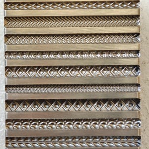 May include: A rectangular metal mold with various decorative border designs. The mold features horizontal rows with different patterns, including chevrons, diamonds, and floral motifs. The metal has a silver-gray color.