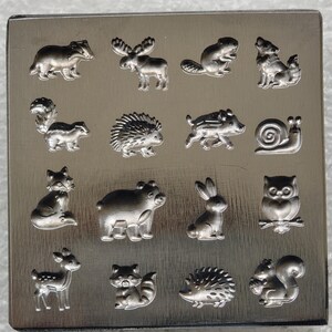 Puede incluir: Una placa metálica cuadrada de tono plateado con dieciséis figuras de animales en relieve dispuestas en una cuadrícula de cuatro por cuatro. Los animales incluyen un tejón, alce, castor, lobo, mofeta, erizo, jabalí, caracol, zorro, oso, conejo, búho, ciervo, mapache, erizo y ardilla.