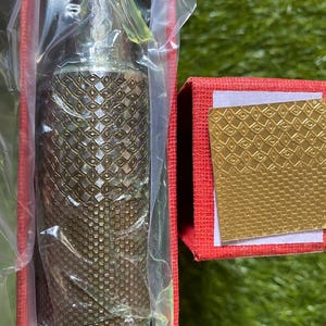 Może przedstawiać: Ozdobny flakon perfum w kolorze brązu ze skomplikowanym wzorem diamentów, zapakowany w plastik. Flakon znajduje się w czerwonym pudełku ze złotym akcentem.