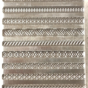 Puede incluir: Un sello metálico rectangular con varios diseños de bordes decorativos. El sello presenta una serie de patrones en relieve, incluyendo espirales, puntos, diamantes y texturas de cuerda. El metal tiene un acabado cepillado.