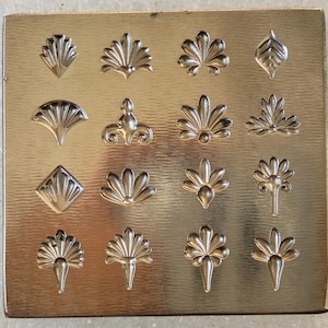 Puede incluir: Una lámina de metal dorada con una serie de diseños florales y geométricos en relieve. Los diseños incluyen hojas estilizadas, formas de abanico y patrones florales simétricos. Los elementos en relieve están realzados y crean una superficie texturizada.