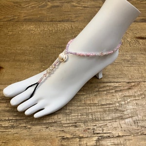 May include: A pink and white beaded foot jewelry with a seashell charm. The jewelry is worn on a white mannequin foot.