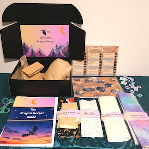 May include: A craft kit in a black box featuring a mountain and castle illustration with the text Welcome, Dragon Keeper. Includes a Dragon Keeper Guide, fabric bundles labelled Treasure Bag, Eggs, Wadding, and Dragon Fabric, plus a reward chart.