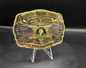 Puede incluir: Una hebilla de cinturón decorativa de metal con las palabras "Franklin County Sheriff" y "Honor, Integrity, Trust" en letras doradas. La hebilla presenta una estrella dorada con un centro verde y el texto "Established 1883".