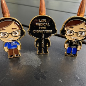 May include: Three gold-coloured pins featuring a cartoon character wearing a headset and glasses. The middle pin has the text "LAW MEDICAL FIRE DISPATCHER" on the back.