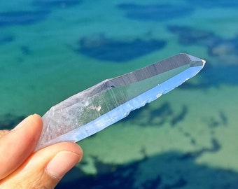 May include: A clear quartz crystal point with a sharp tip and a smooth, polished surface. The crystal is held in a hand against a blurred background of blue water.