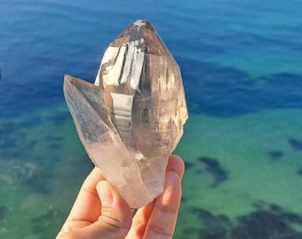 May include: A large, clear quartz crystal with a smoky brown point. The crystal is held in a hand against a background of blue and green water.