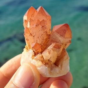 Könnte beinhalten: Ein Cluster aus rosa und orangefarbenen Quarzkristallen mit einer rauen, natürlichen Textur. Die Kristalle sind in einem Cluster angeordnet, wobei einige der Kristalle nach oben zeigen. Die Kristalle sind vor einem Hintergrund aus blauem Wasser platziert.