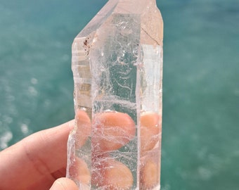 May include: A clear quartz crystal point with a natural, rough texture. The crystal is held in a hand against a blue water background.