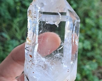 May include: A clear quartz crystal with a double termination and inclusions of black and red minerals. The crystal is approximately 4 inches tall and has a natural, rough texture.
