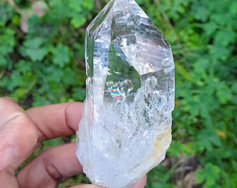 May include: A large, clear quartz crystal with a double termination and rainbow iridescence. The crystal is held in a hand, and the background is blurred.