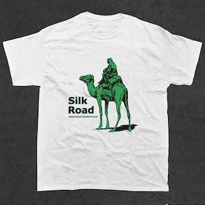 Pode incluir: Camiseta branca com uma ilustração verde de uma pessoa montando um camelo. O texto "Silk Road" está impresso em verde abaixo da imagem, com as palavras "Anonymous Marketplace" em verde menor abaixo.