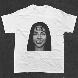 May include: White t-shirt with a black and white image of a woman's face. The text "FREE THUG" is printed across her forehead.