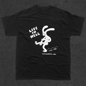 May include: Black t-shirt featuring a white graphic of a rabbit kicking a small object. The text "Life in Hell" is above the rabbit and "Matt Groening ©1984" is below the rabbit.