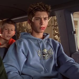 Op de afbeelding: Een jonge man draagt een lichtblauw hoodie met een schedel en vlammen afbeelding op de voorkant. Hij zit in een auto met een andere jonge man op de passagiersstoel.