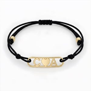 Può includere: Un braccialetto dorato con un cordino nero. Il braccialetto presenta le lettere "C", un cuore e "A" in un design rettangolare. Il cordino ha perline dorate a ciascuna estremità.