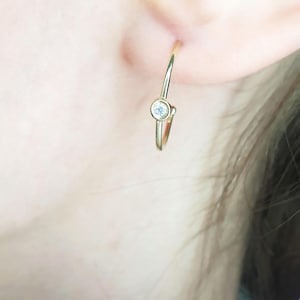 May include: A gold hoop earring with a small, round, clear gemstone set in the center. The earring is worn on the ear, with the post inserted through the earlobe. The earring is simple and elegant.
