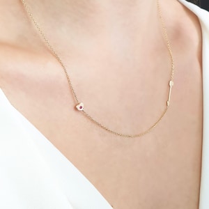 Può includere: Una collana d'oro con un piccolo ciondolo a forma di cuore e un ciondolo a forma di freccia. Il ciondolo a forma di cuore ha una pietra preziosa rossa al centro.