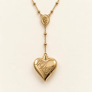 Puede incluir: Un collar de tono dorado con una cadena delicada de pequeñas cuentas esféricas y un dije central circular con la figura de un querubín en relieve. Un colgante liso en forma de corazón cuelga de la cadena, grabado con las letras cursivas LDR.