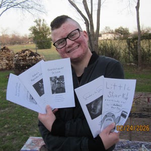 May include: A person holding several small books with white covers. The titles include "Deathmetalist In Love" and "Little Shark!" The books have black and white illustrations and text. The background shows a park setting with trees and a bench.