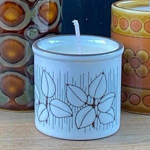 May include: A light blue ceramic candle holder with a floral design in brown and white. The candle holder is filled with white wax and has a pink wick. Two other decorative ceramic vases are in the background.
