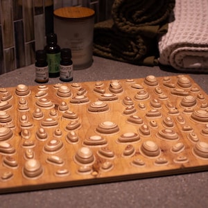 Op de afbeelding: Een houten badmat met een uniek gelaagd ontwerp. De mat is gemaakt van lichtbruin hout en heeft talrijke verhoogde, cirkelvormige vormen van verschillende afmetingen. Het ontwerp lijkt op een topografische kaart. Verschillende essentiële olieflessen en opgerolde handdoeken staan op de achtergrond.