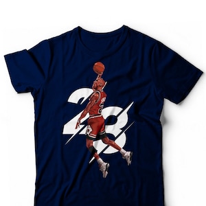 Könnte beinhalten: Marineblaues T-Shirt mit einer Grafik eines Basketballspielers im Sprung, kurz vor dem Wurf. Der Spieler trägt eine rot-weiße Uniform. Die Nummer "23" ist weiß dargestellt.