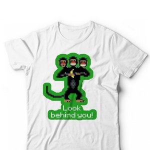 May include: White t-shirt with a green pixelated image of a monkey with three heads. The monkey is holding a banana and the text "Look behind you!" is below the image.