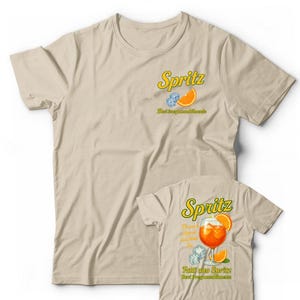 Peut inclure: T-shirt beige avec un motif Spritz. Le devant présente le mot "Spritz" en jaune, une tranche d'orange et des glaçons. Le dos du t-shirt affiche un cocktail Spritz dans un verre avec du texte.