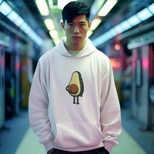 Può includere: Una felpa con cappuccio bianca con un design di avocado a cartone animato. L'avocado è verde e marrone e ha un viso sorridente. La persona che indossa la felpa è in piedi in una stazione della metropolitana.