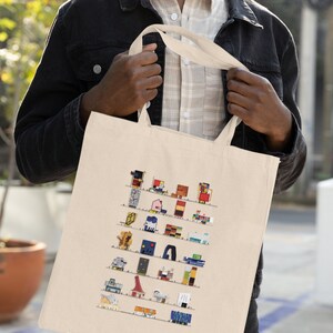 Op de afbeelding: Een witte tote bag met een kleurrijk ontwerp van verschillende gebouwen. De tas heeft twee handvatten en wordt door een persoon vastgehouden.