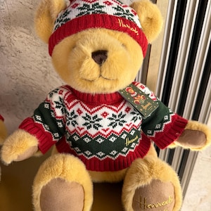 May include: A soft teddy bear wearing a red and white knitted hat with a bobble and a matching sweater. The sweater features a green and white snowflake design. The bear has the Harrods logo on its foot and a tag.