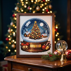 Puede incluir: Un patrón de punto de cruz enmarcado que representa una bola de nieve con un árbol de Navidad decorado, dos casas pequeñas y un paisaje nevado. La base de la bola presenta un lazo rojo, campanas doradas y un bastón de caramelo, en un marco.