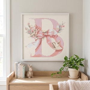 Puede incluir: Una pieza de arte en punto de cruz con marco cuadrado que presenta una letra B grande de color rosa decorada con un arreglo floral, un lazo rosa prominente y una pequeña ilustración de un conejo blanco sobre un fondo color crema.