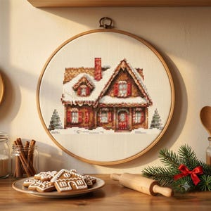 Puede incluir: Un bastidor de bordado circular de madera que contiene un patrón de punto de cruz de una casa marrón con chimenea y contraventanas rojas, cubierta de nieve blanca. La casa está flanqueada por dos pequeños pinos verdes sobre tela crema.
