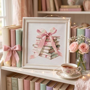 May include: A framed cross-stitch artwork depicting a stack of books tied with a large pink ribbon. The piece is displayed on a shelf alongside pastel-colored books, a small glass vase with two pink roses, and a floral teacup on a saucer.