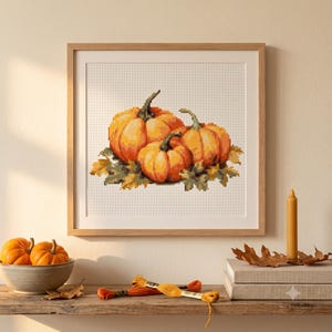 May include: A square framed cross-stitch pattern featuring three orange pumpkins nestled among autumn leaves. The design is rendered in a pixelated style with visible grid lines, showcasing shades of orange, green, and brown on a white background.