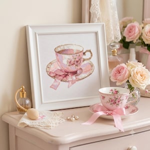 Peut inclure: Une broderie au point de croix encadrée représentant une tasse à thé rose avec des roses et un ruban, exposée à côté d'une tasse en porcelaine assortie avec un nœud en satin rose, un vaporisateur de parfum, un napperon et un vase de roses.