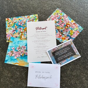 May include: A collection of stationery, including cards and an envelope, is displayed on a dark surface. The cards showcase floral watercolour designs in pink, blue, and yellow. The envelope and a card display the text "Blütenpost".