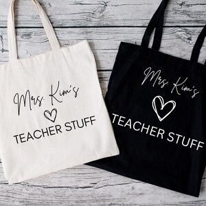May include: Two tote bags, one cream and one black, with white text. The text on each bag reads "Mrs. Kim's TEACHER STUFF" with a heart symbol. The bags have long handles and are made of a sturdy fabric.