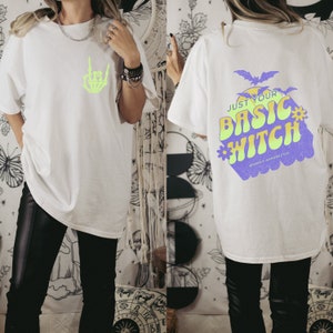 May include: White t-shirt with a green skeleton hand graphic on the front and a purple graphic that says "Just Your Basic Witch" on the back. The graphic also includes a flower and bats.